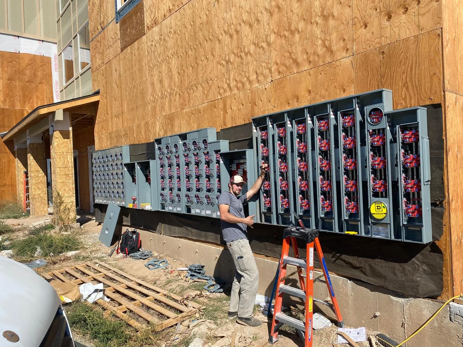 A man at a construction site is installing electrical meters on a building. He is holding one meter in his hand, standing next to an orange ladder, with construction materials and tools scattered around.