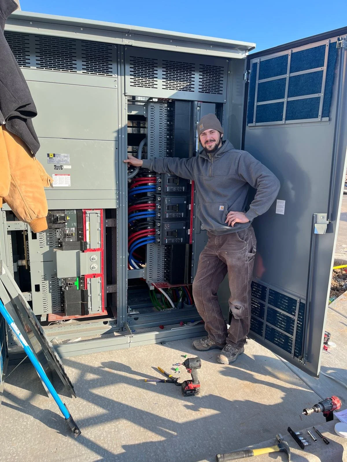 A man in work clothes and a beanie standing next to an open electrical control panel with wires and components, outdoors on a sunny day, with tools on the ground.