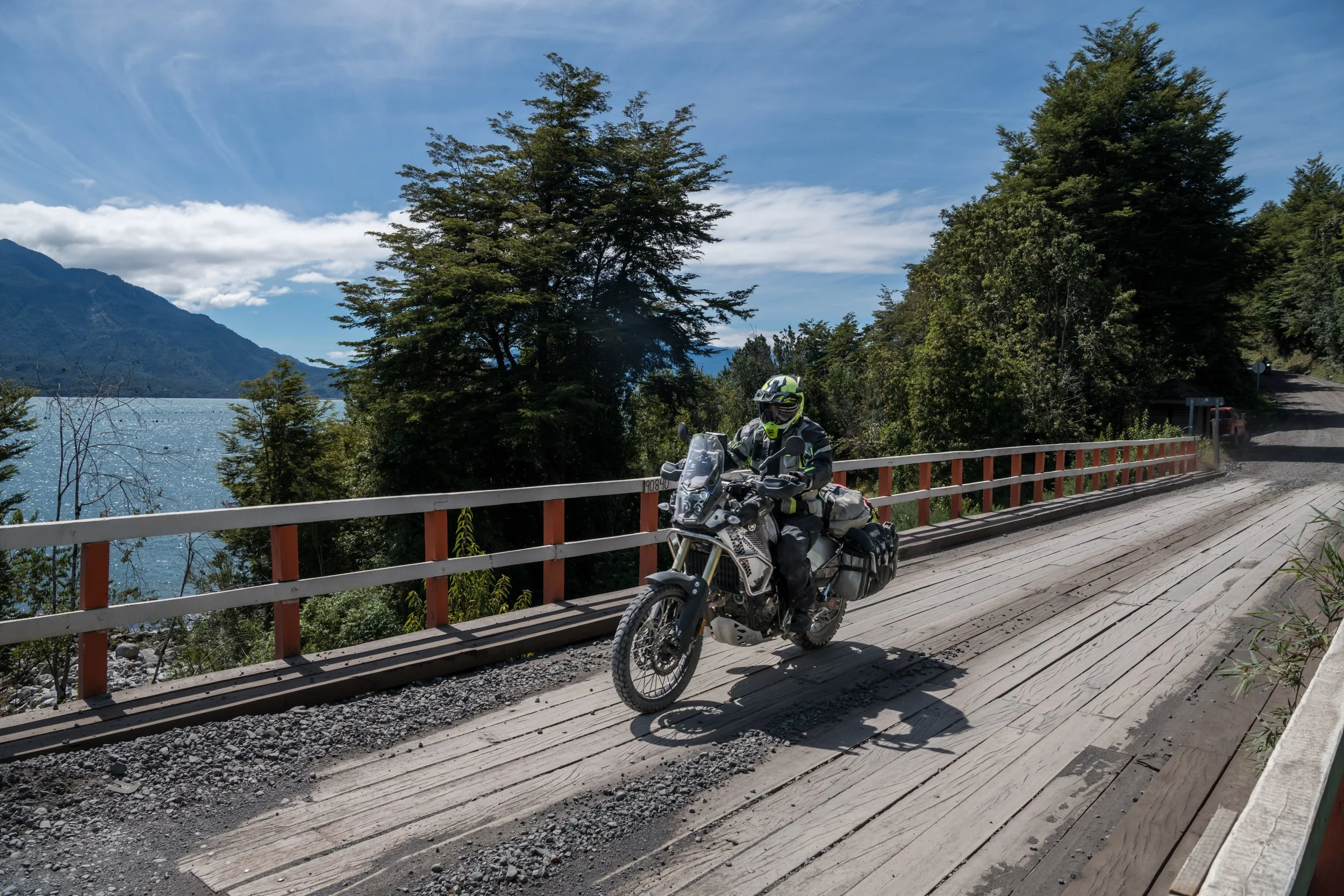 Motorcycle rider wearing a helmet and protective gear riding along a scenic mountain road with trees, a body of water, and mountains in the background.