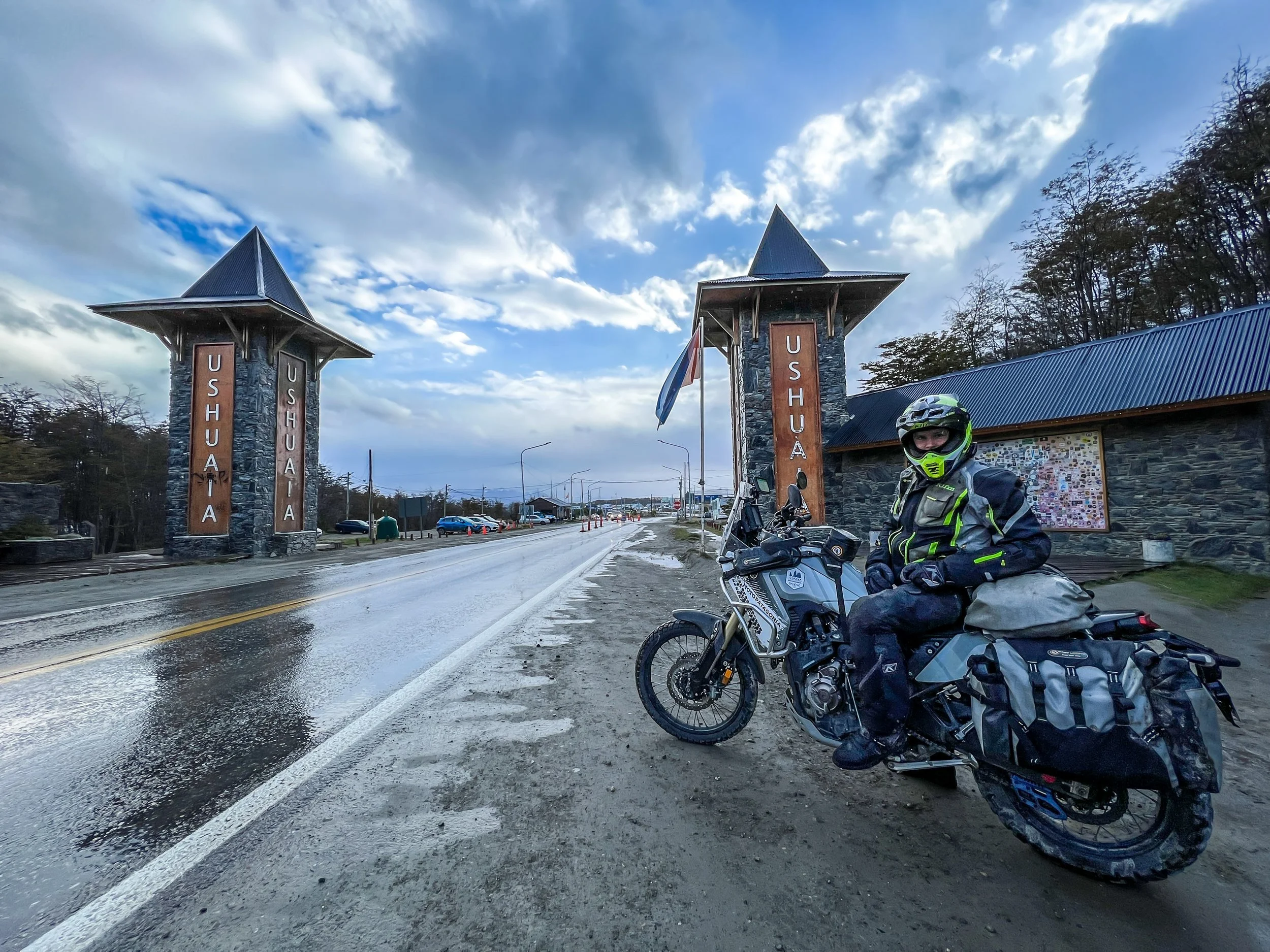 A man in motorcycle gear sitting on a touring motorcycle near the border crossing entrance of Ushuaia, Argentina, with two stone towers and flags, a cloudy sky, and a wet road.
