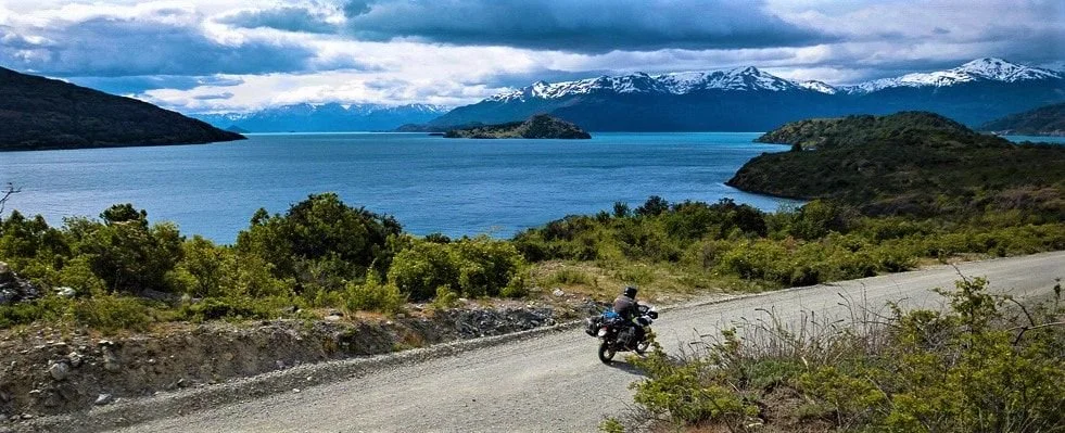 Motorcyclist riding on a dirt road near a large lake, with mountains in the background and snow on their peaks.