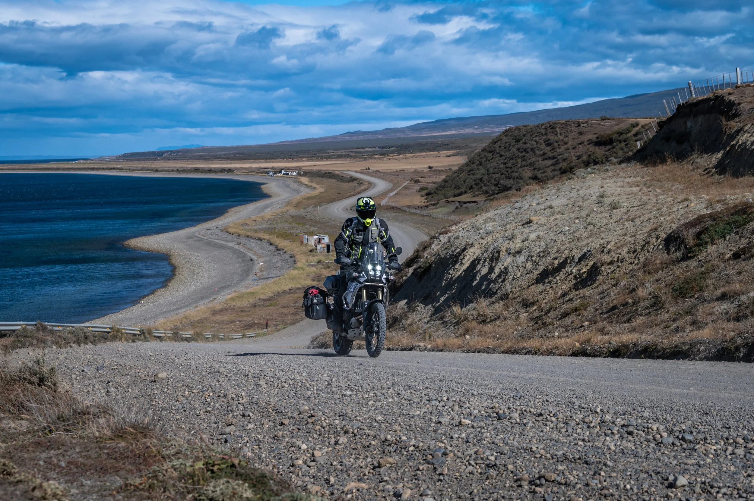 Motorcycle with rider on a gravel road along a body of water under a partly cloudy sky in a rural landscape.