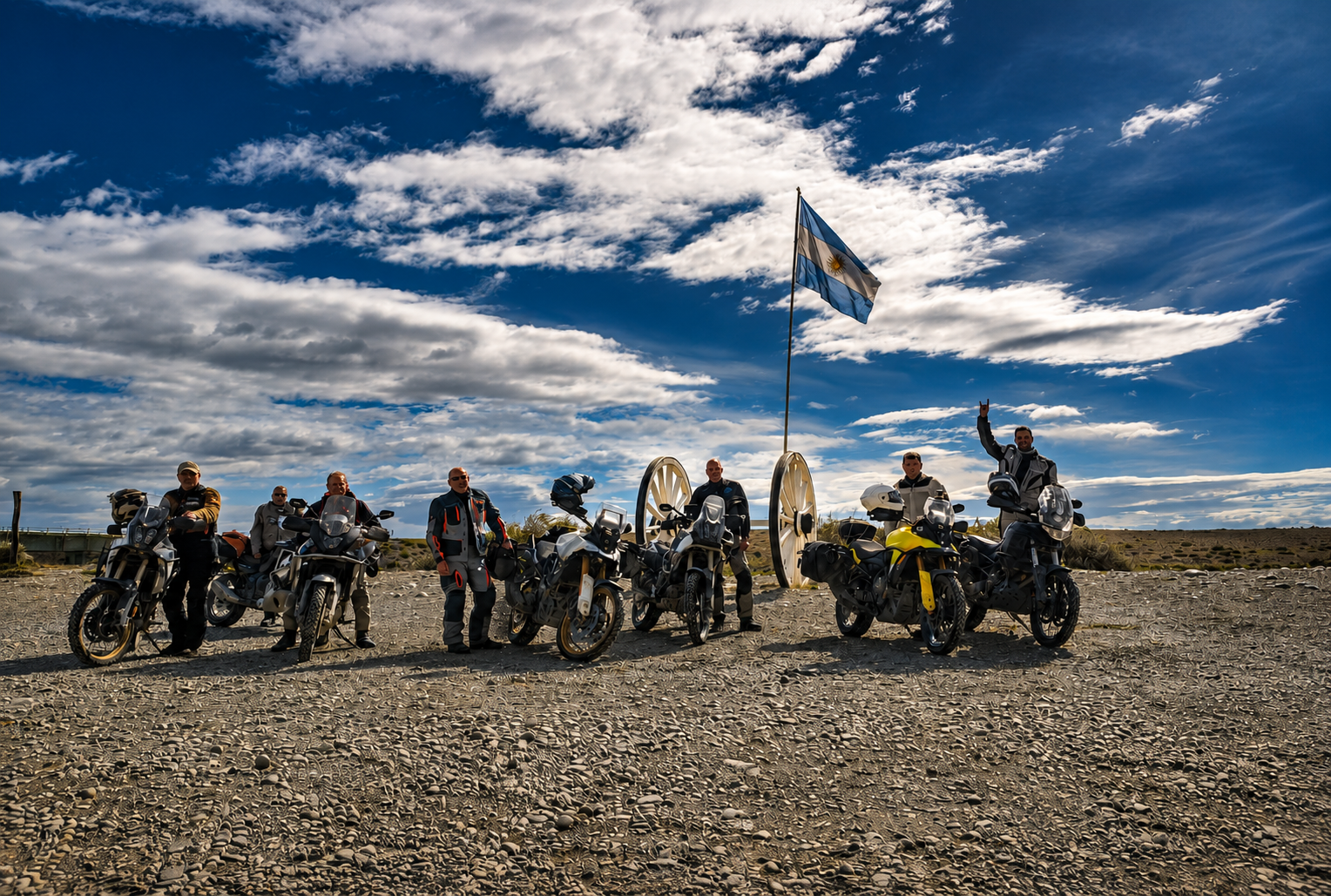 Group of seven motorcyclists in riding gear standing beside their motorcycles on rocky terrain under a partly cloudy sky, with an Argentine flag on a pole and an old cannon in the background.