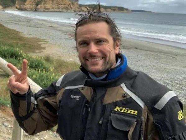 A man smiling and making a peace sign with his hand on a beach with cliffs and ocean in the background.