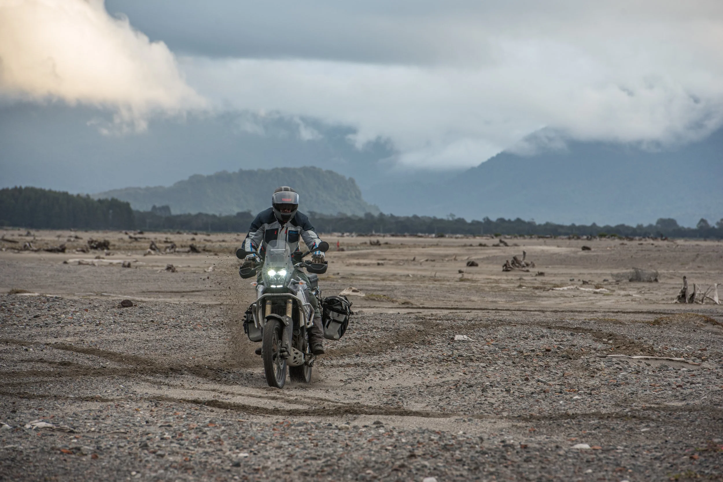 A person riding a motorcycle on a dirt path in a desolate landscape with mountains and cloudy sky in the background.