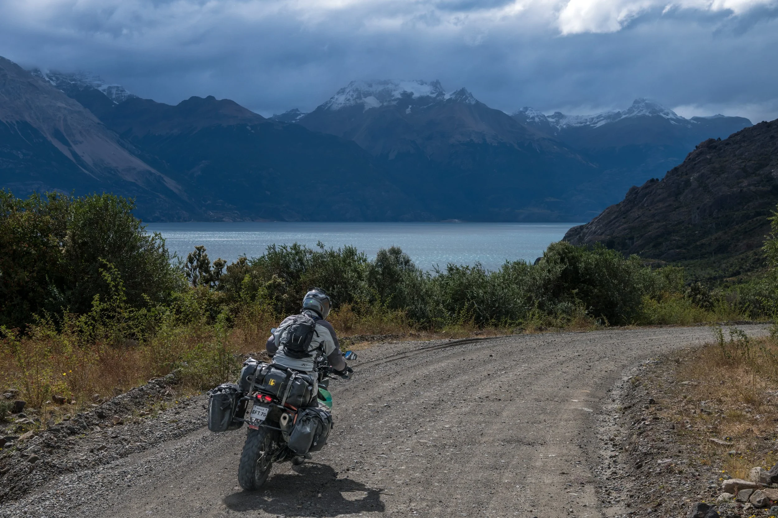 A person riding a motorcycle on a dirt road with a scenic view of a lake and towering snow-capped mountains in the background, under cloudy skies.
