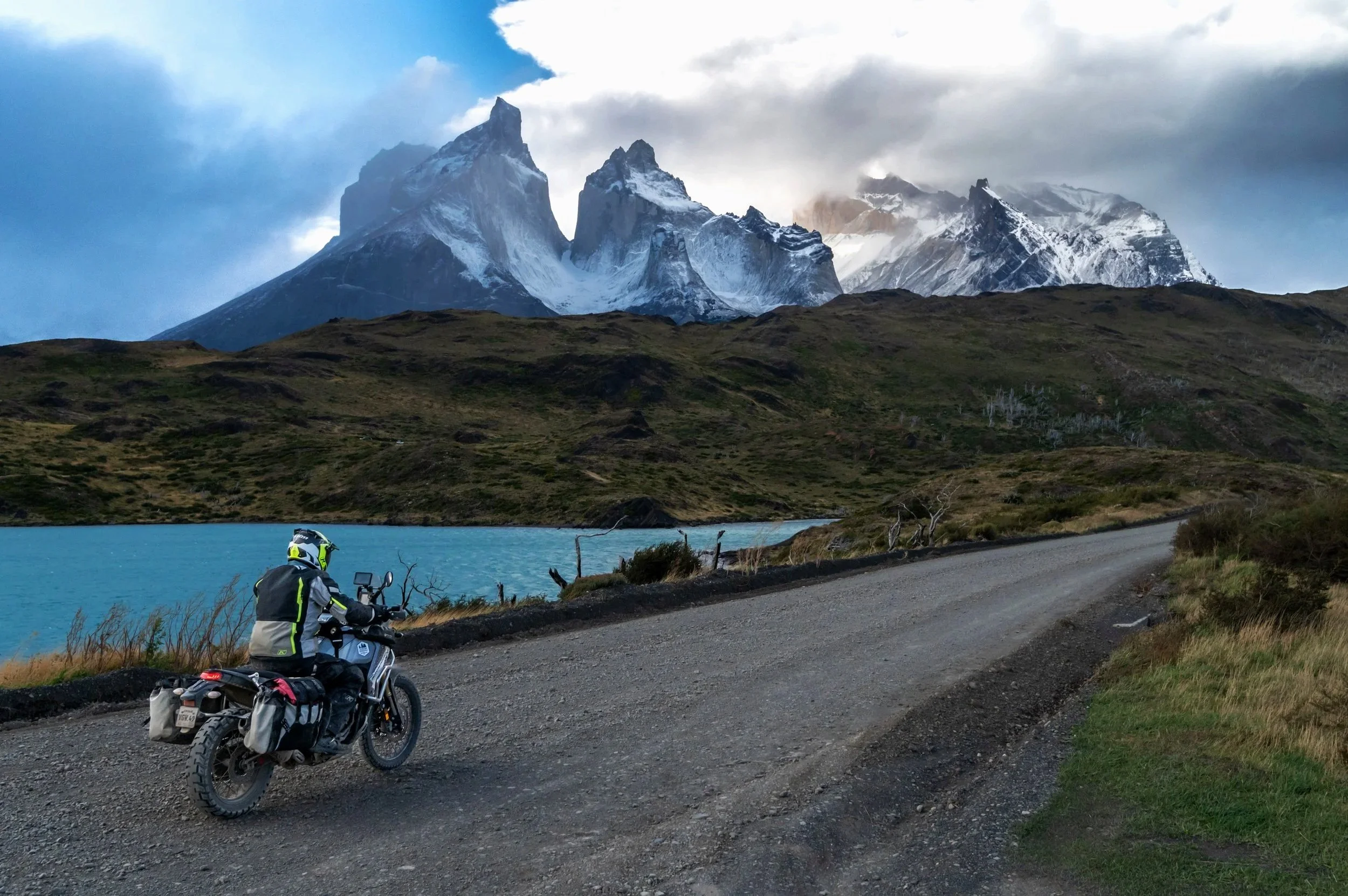 A motorcyclist wearing a helmet and riding gear is parked on a gravel roadside near a turquoise lake, with snow-capped mountains and a cloudy sky in the background.