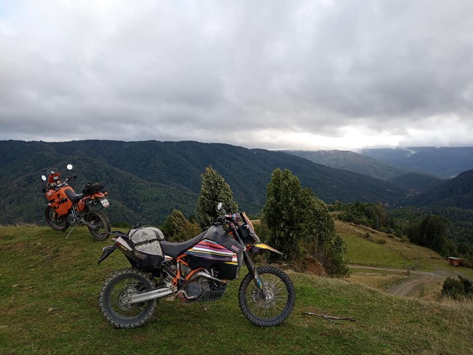 Two off-road motorcycles parked on a grassy hill with a mountainous landscape and cloudy sky in the background.