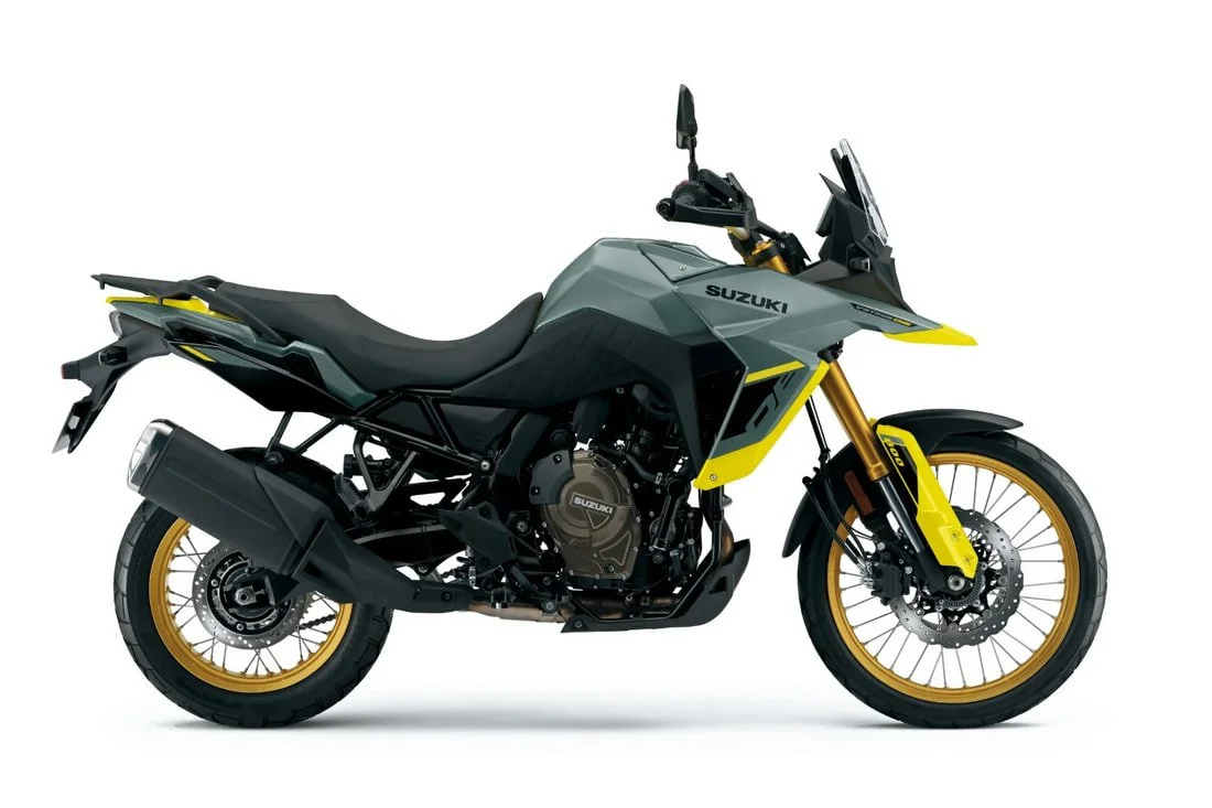 A modern Suzuki adventure motorcycle with a matte gray body, yellow accents, gold front forks, spoked wheels, and a rugged design suitable for off-road riding.