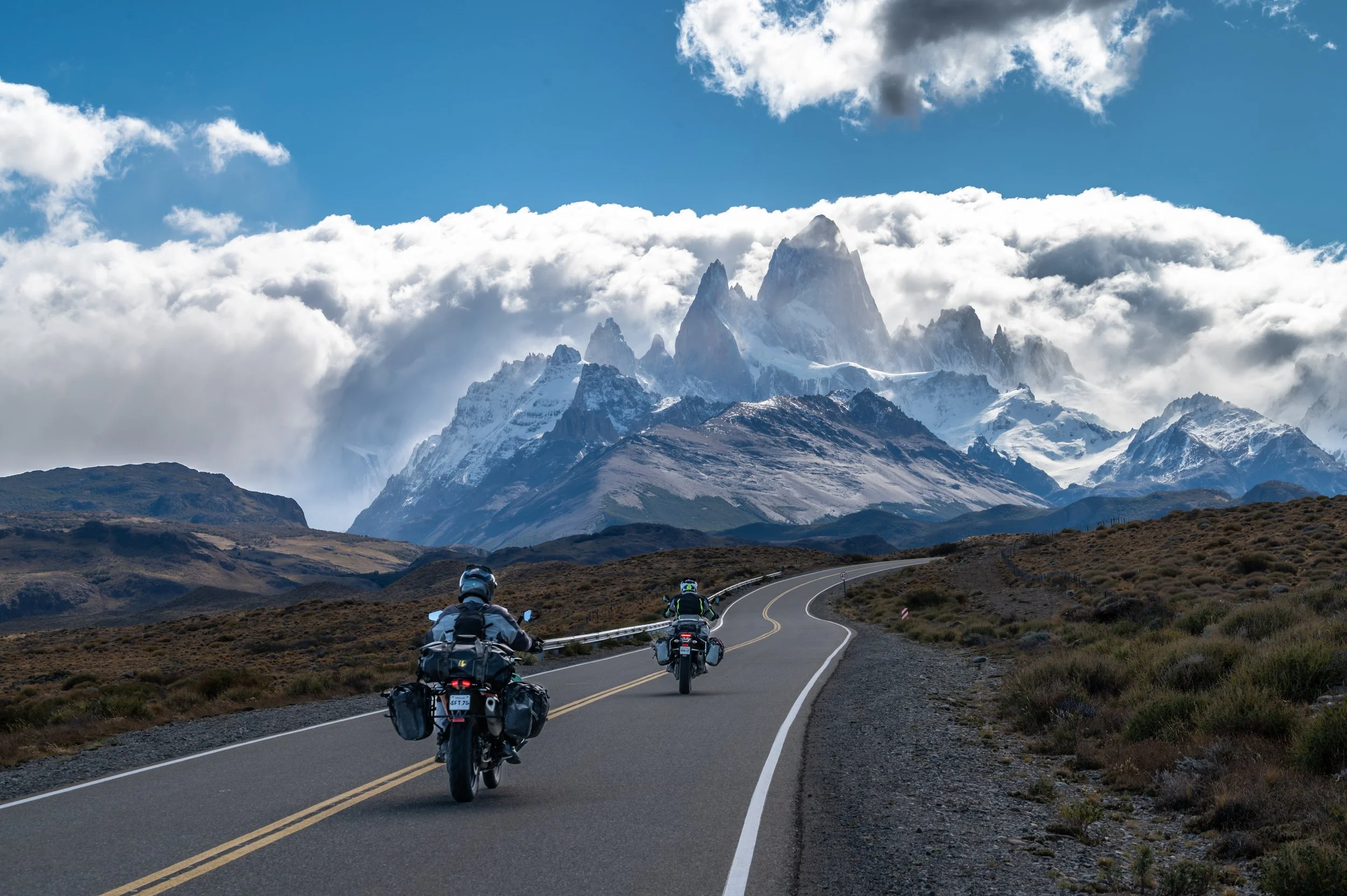 Four motorcyclists riding on a winding mountain road with snow-capped peaks and clouds in the background.