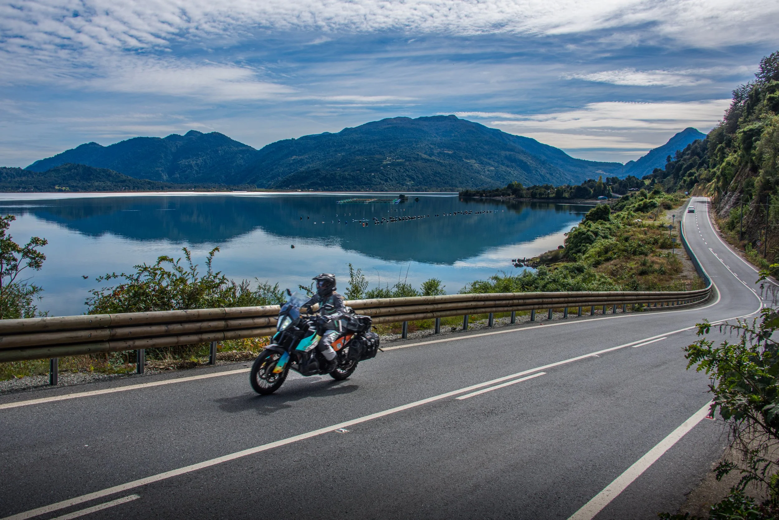 A motorcycle riding on a winding mountain road next to a lake with mountains in the background and a mostly cloudy sky.