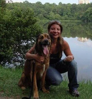 A woman kneeling beside a large brown dog by a lake, with trees and greenery in the background.