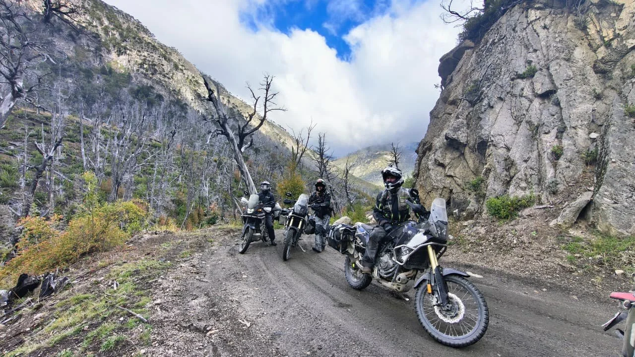 Four motorcyclists wearing helmets and gear stop on a dirt mountain trail surrounded by rocky cliffs and leafless trees with a cloudy sky overhead.