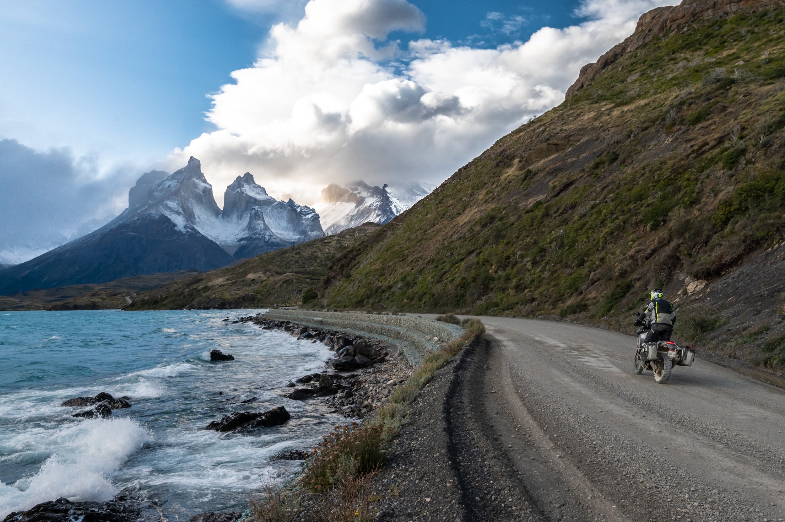 A scenic landscape showing a winding road along a mountain lake with snow-capped peaks in the background, and a motorcycle with a rider wearing a helmet and gear parked on the side of the road.