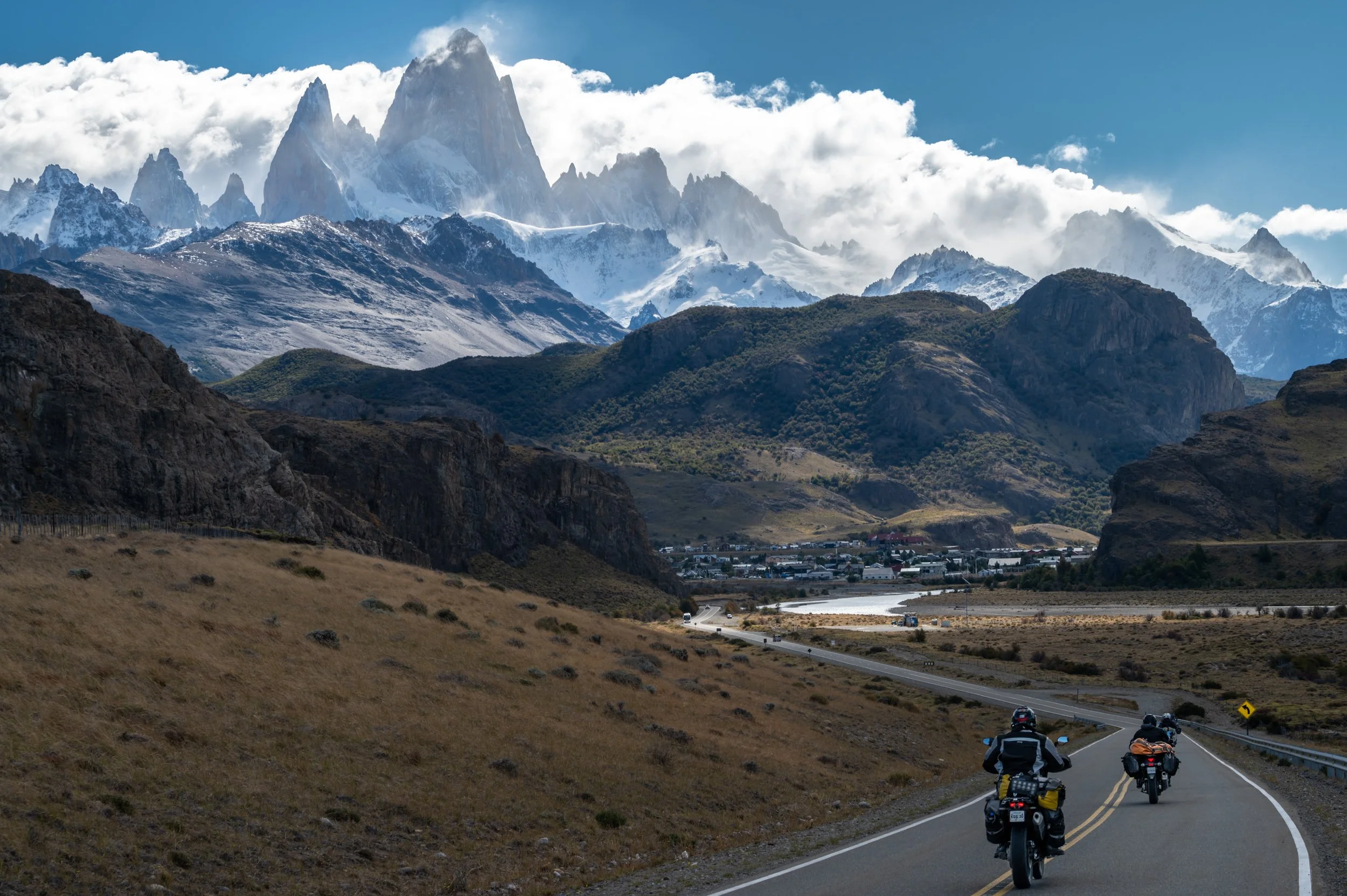 Group of motorcyclists riding on a winding mountain road with snow-capped peaks and rocky terrain in the background.