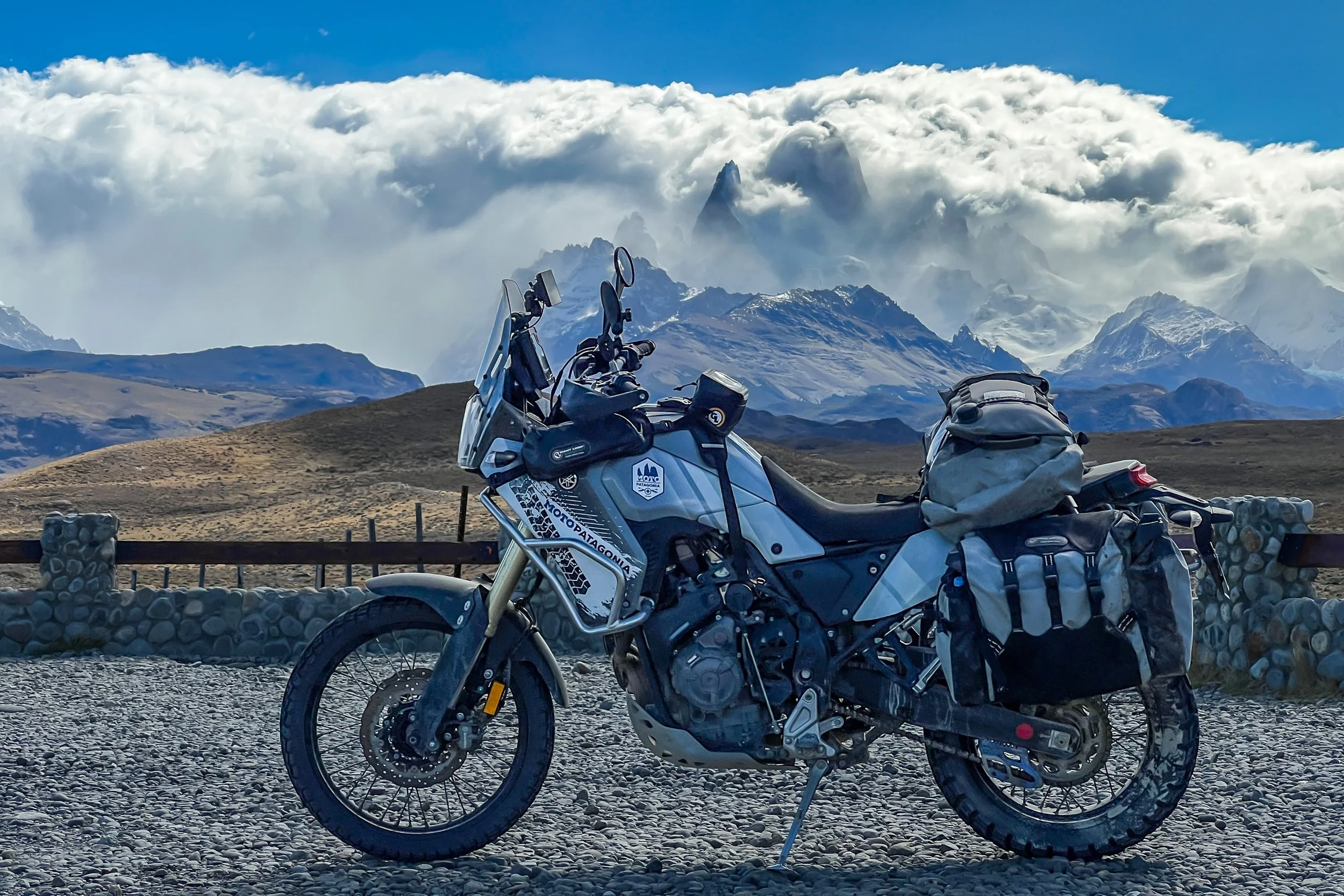 A motorcycle parked on a gravel surface with a mountain range and clouds in the background.
