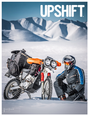 A person in motorcycle gear kneeling next to a bicycle with front bags, set in a snowy mountain landscape with the word UPSHIFT at the top.
