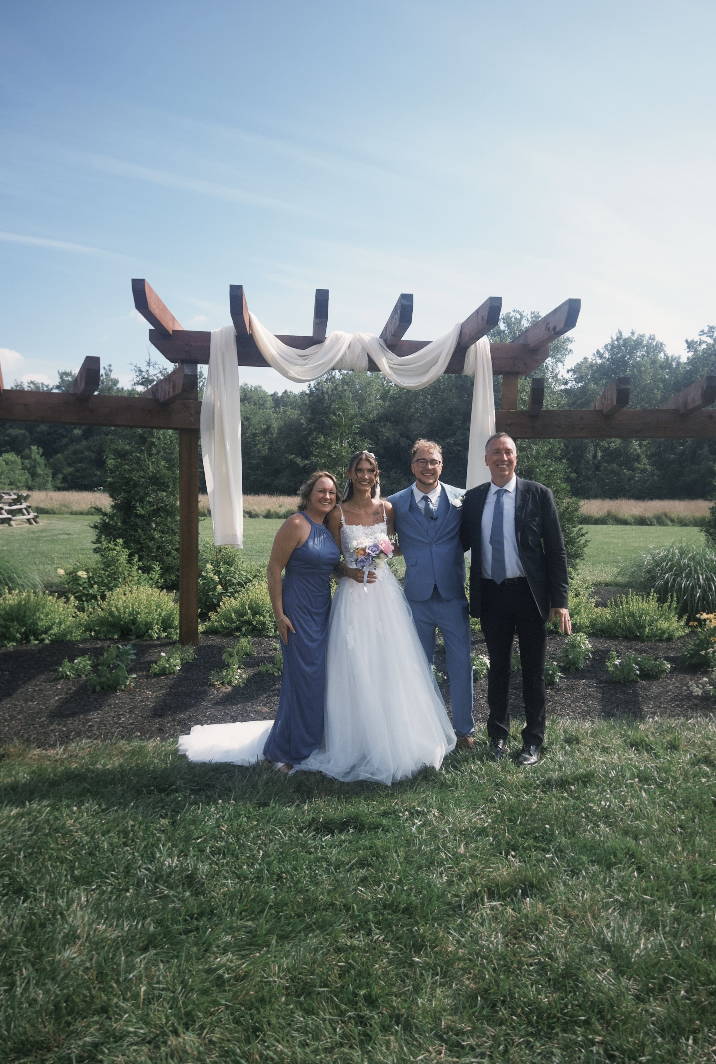 Four people standing outdoors in front of a wooden arch decorated with white fabric at a wedding ceremony. The group includes a bride in a white wedding gown holding a bouquet, a woman in a blue dress, a man in a light blue suit, and a man in a dark suit. There is greenery, trees, and open sky in the background.