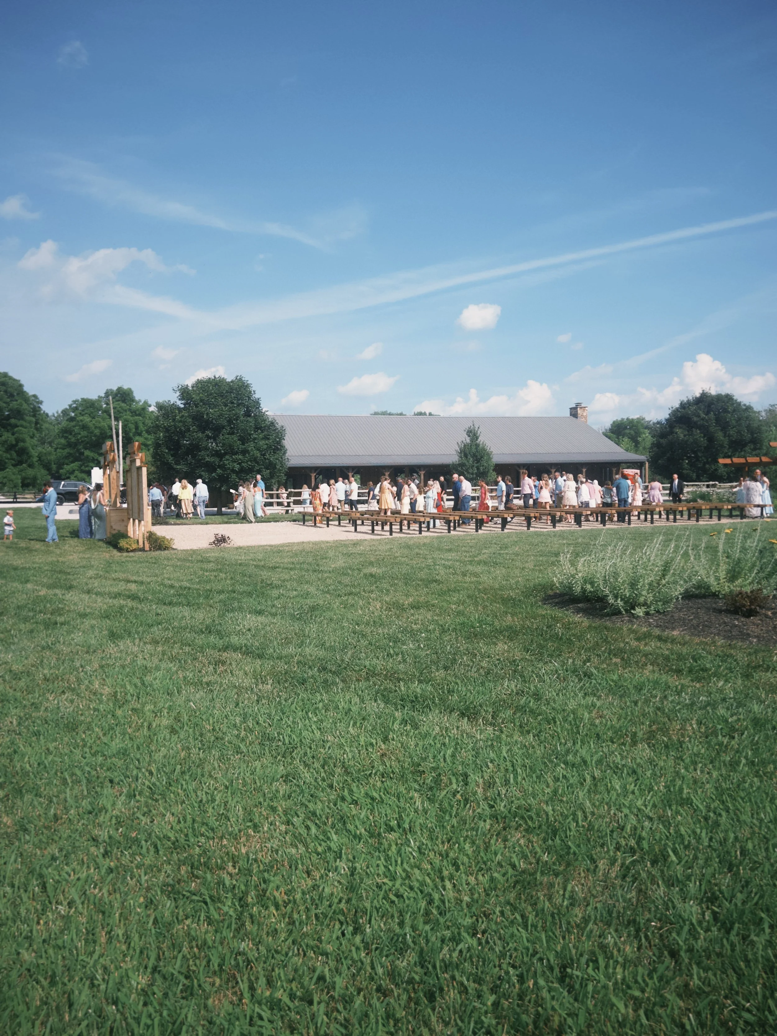 A large group of people gathered outdoors near a building under a blue sky with some clouds and trees in the background, possibly at an event or celebration.