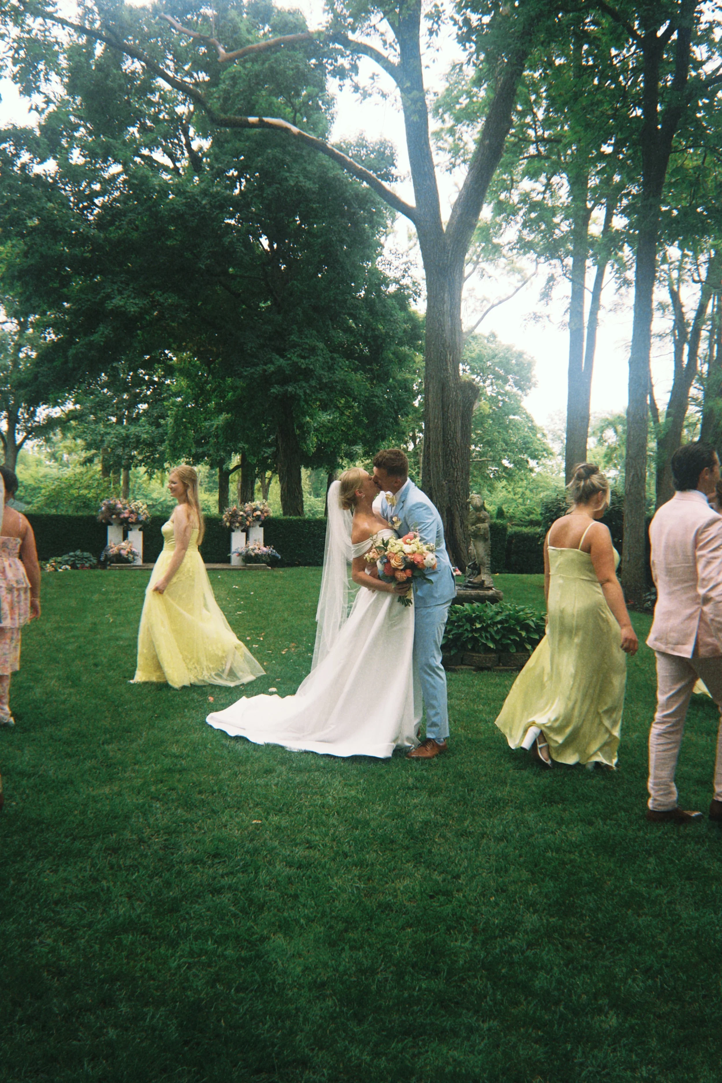 A bride and groom kissing outdoors at a wedding reception, surrounded by guests in colorful dresses and suits, with large trees and floral arrangements in the background.