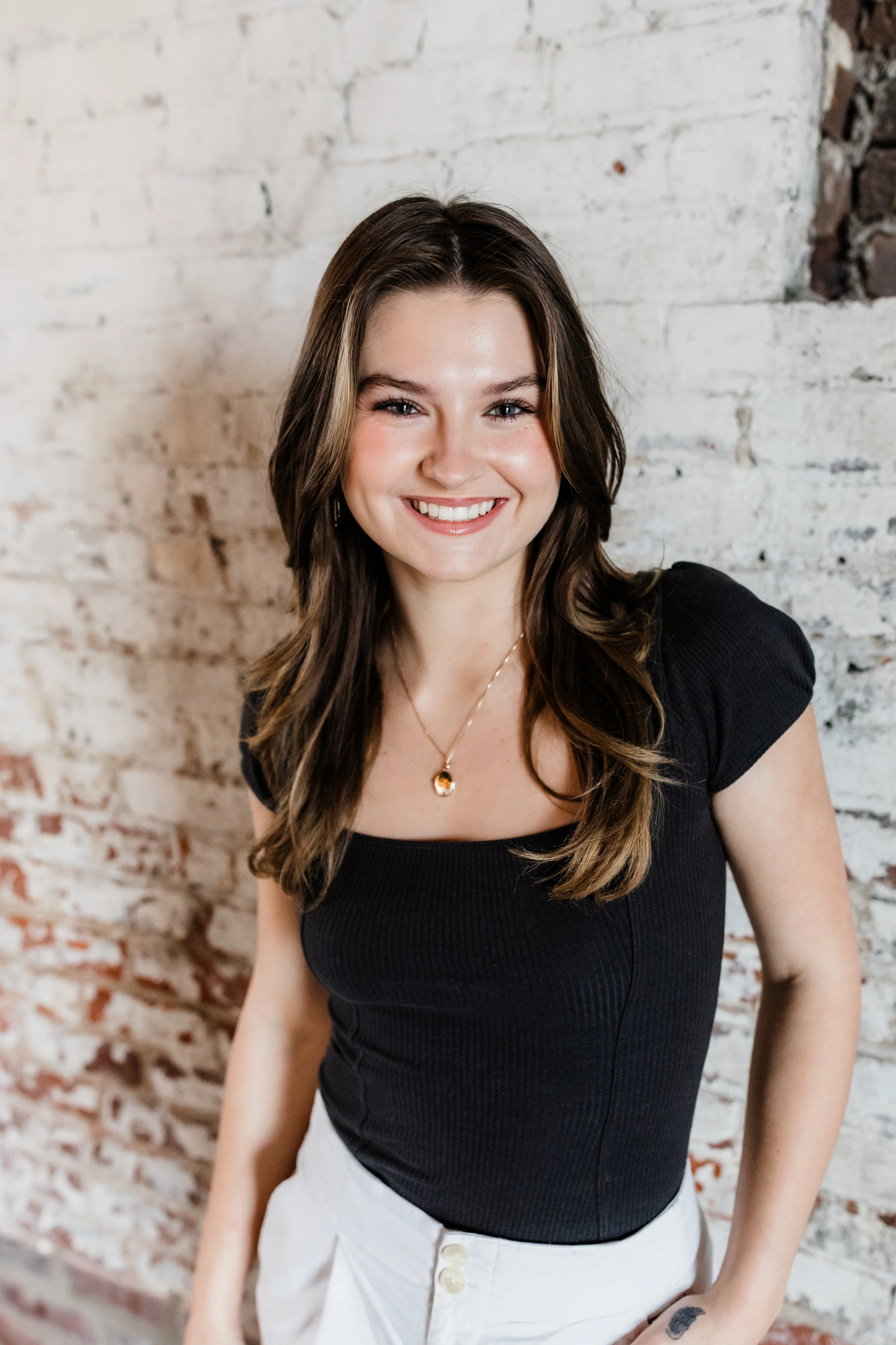 A young woman with long, wavy brown hair and a warm smile stands in front of a white brick wall with some exposed bricks. She is wearing a black top, white pants, and a gold necklace with a pendant.