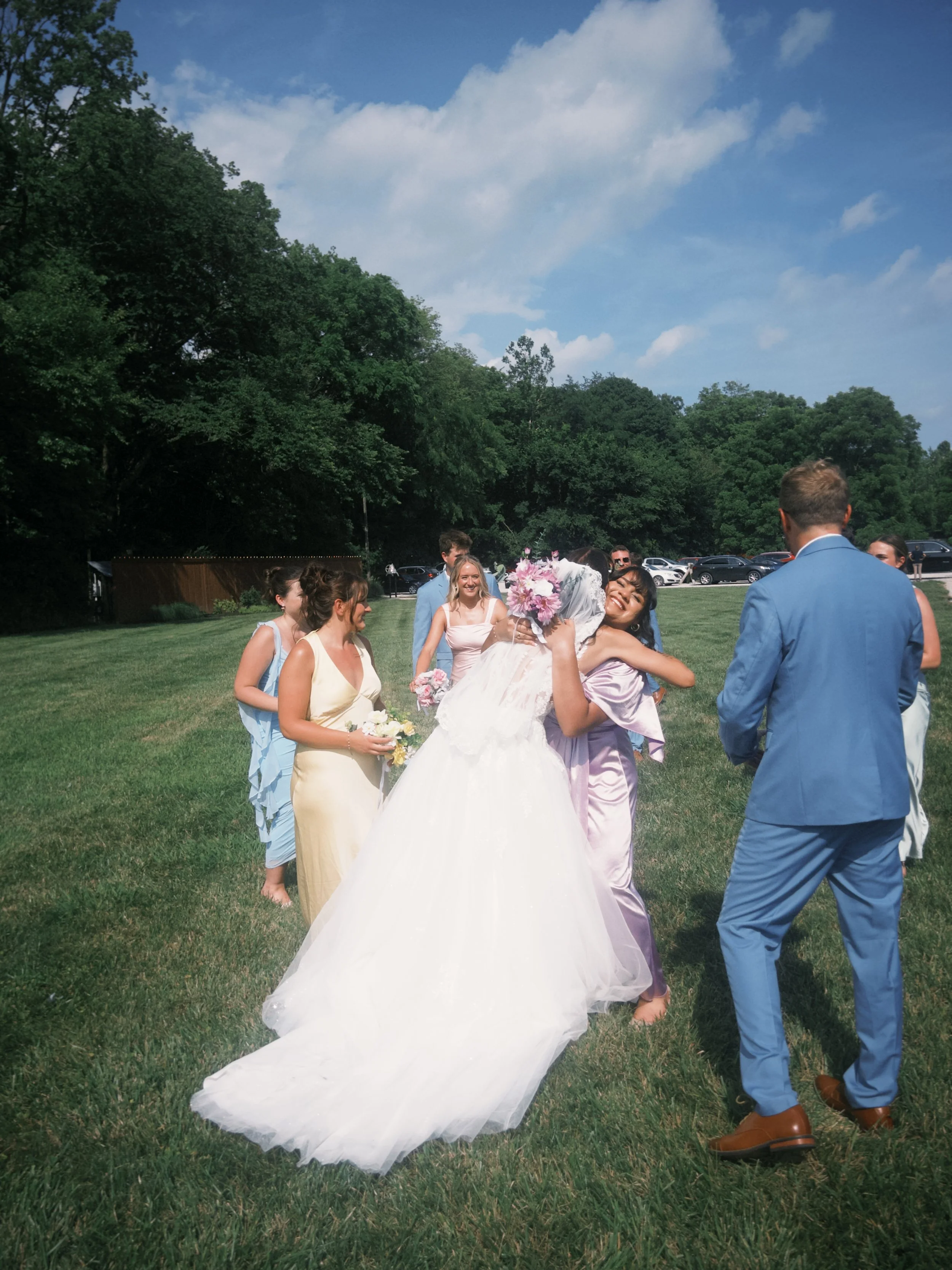 A group of people dressed in formal attire, including a woman in a white wedding gown and a man in a blue suit, gathers outdoors on green grass with trees and cars in the background, celebrating a wedding. The woman is holding a bouquet of flowers, and others surrounding her are smiling and chatting under a partly cloudy sky.