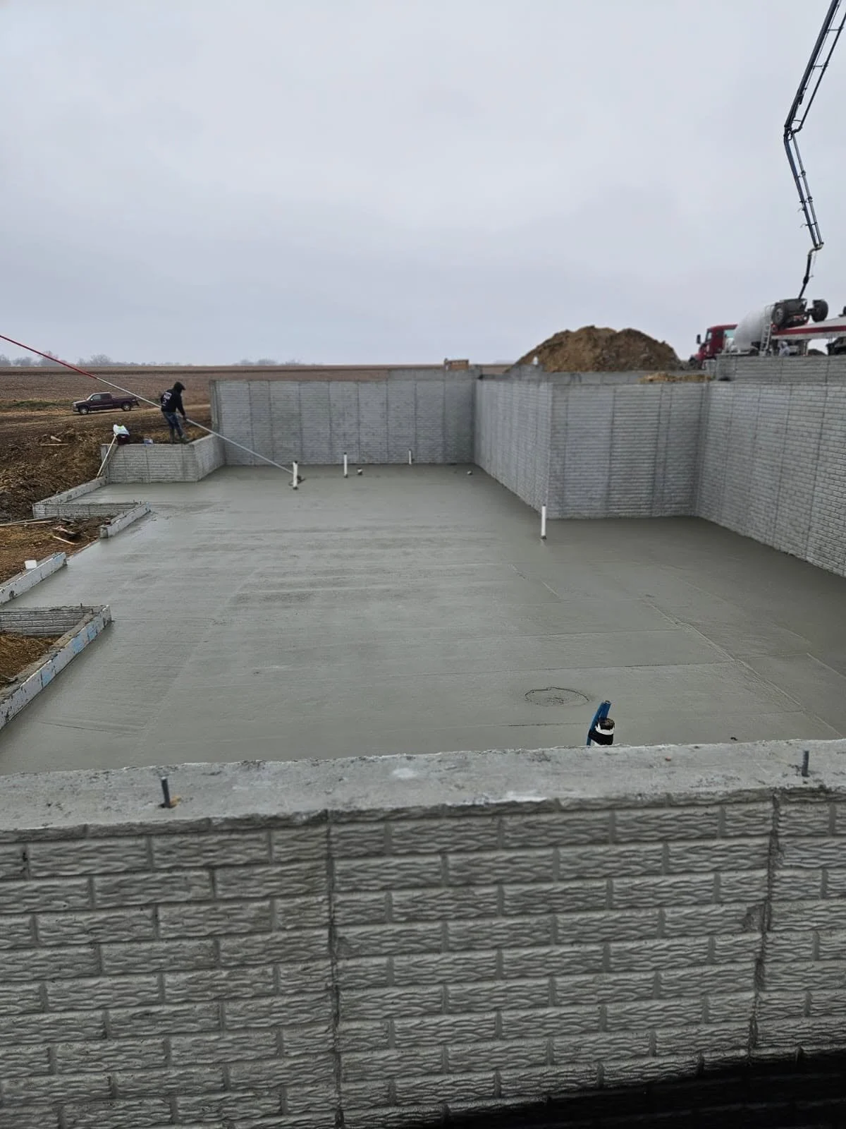 Construction site showing a concrete foundation with workers and a corrugated metal block wall, with construction equipment and an empty field in the background.
