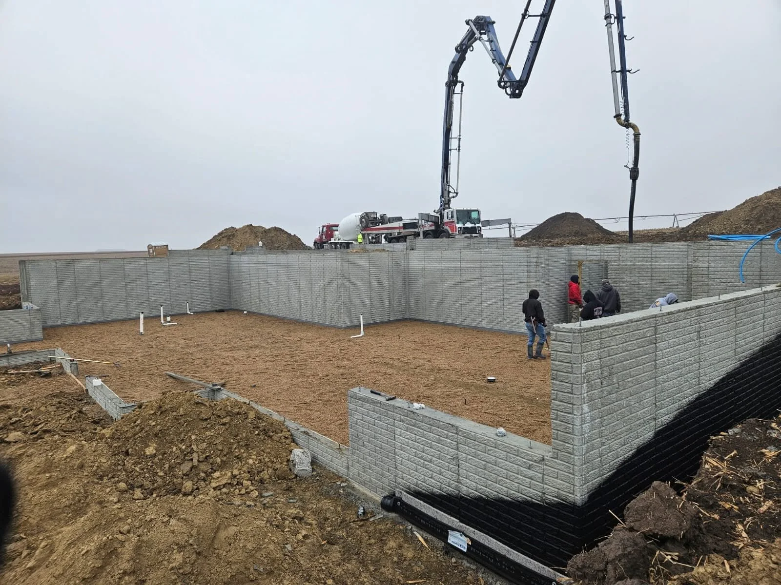 Construction site with workers building a concrete wall, with a large cement mixer truck and concrete pump in the background, and the ground cleared and leveled for further construction.
