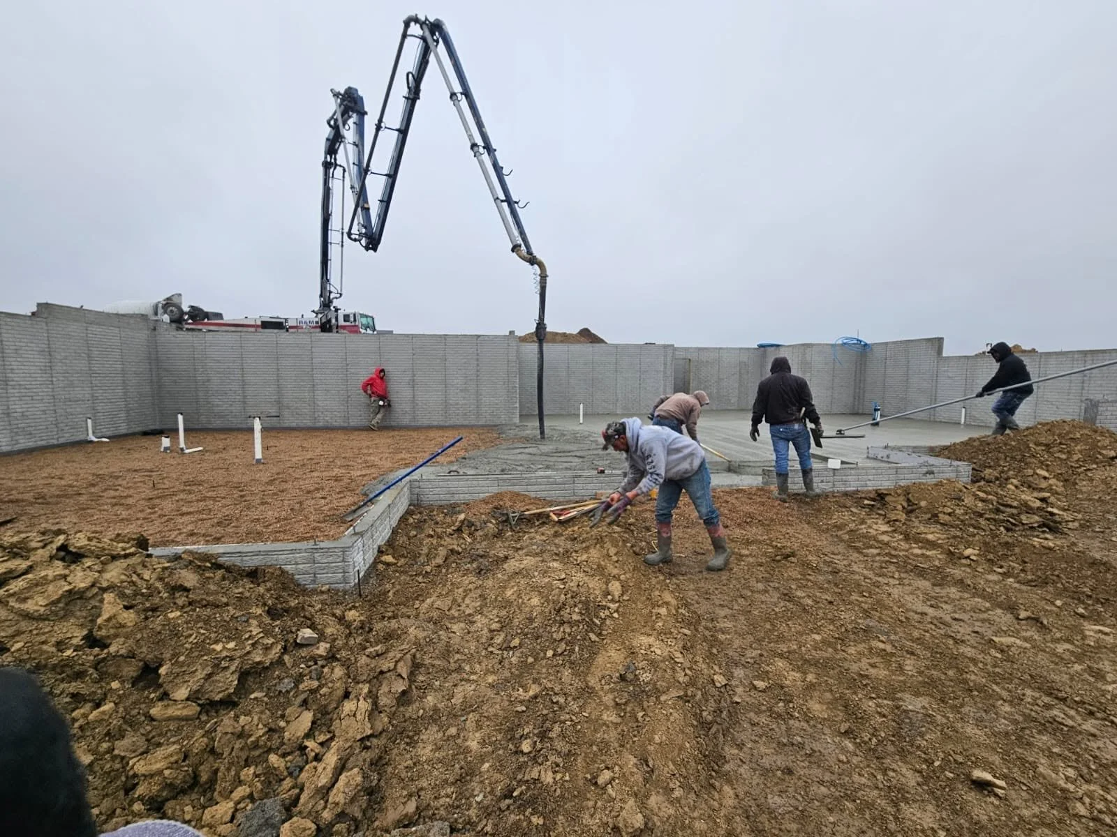 Workers laying bricks and concrete on a construction site surrounded by dirt and a tall fence, with a concrete pump truck in the background.