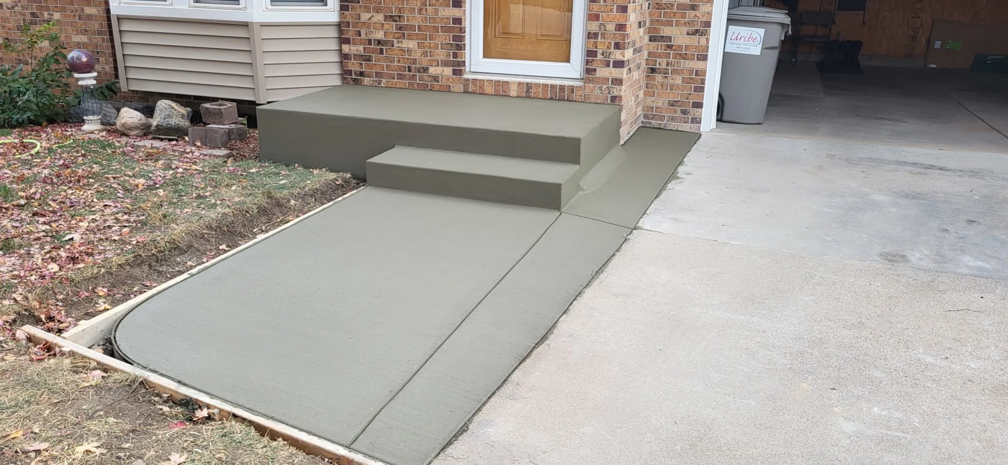 Freshly poured concrete sidewalk and steps at the front entrance of a house, with a brick exterior and a window.