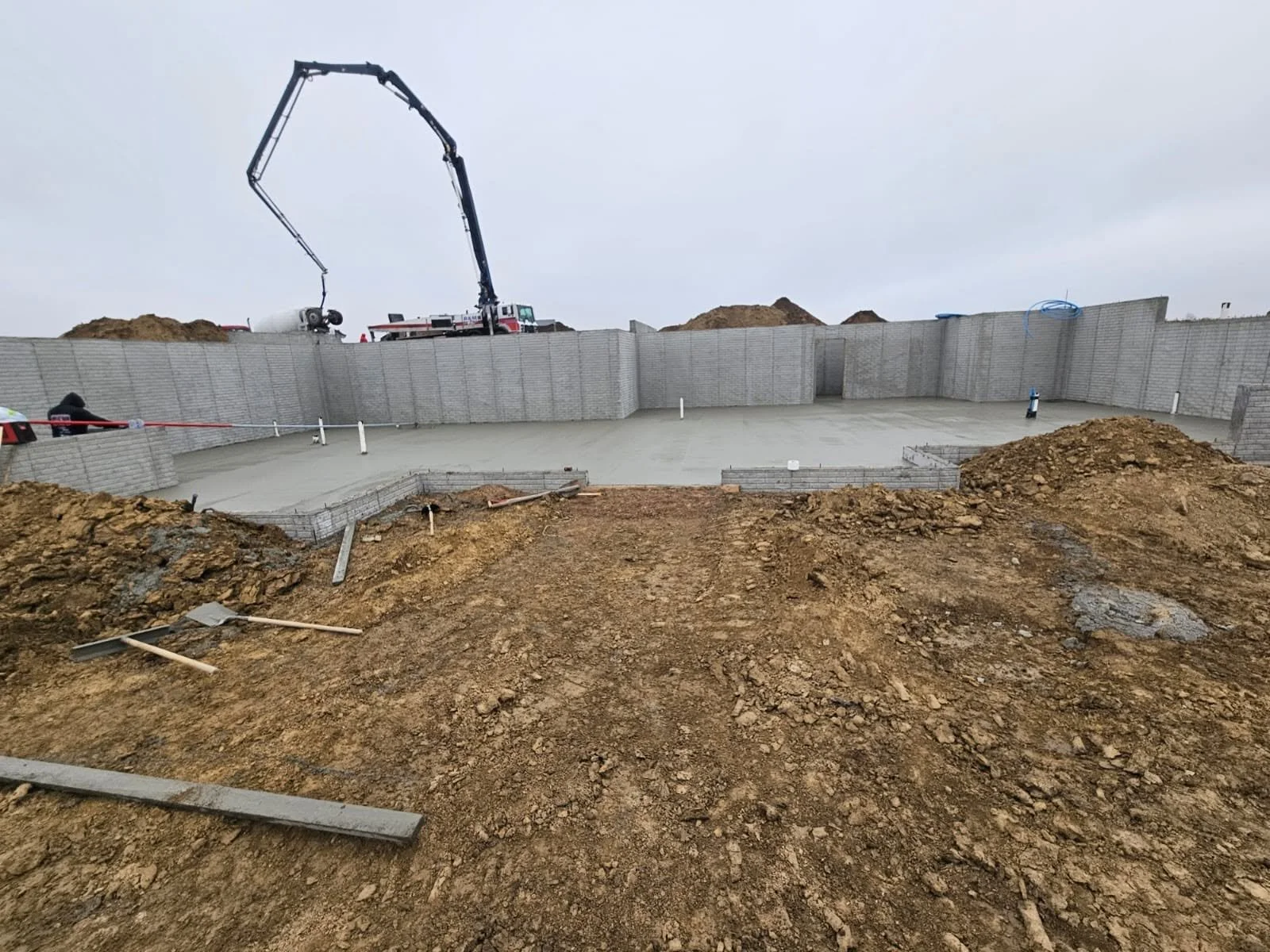Construction site with a partially completed concrete foundation and a crane in the background, surrounded by dirt piles.