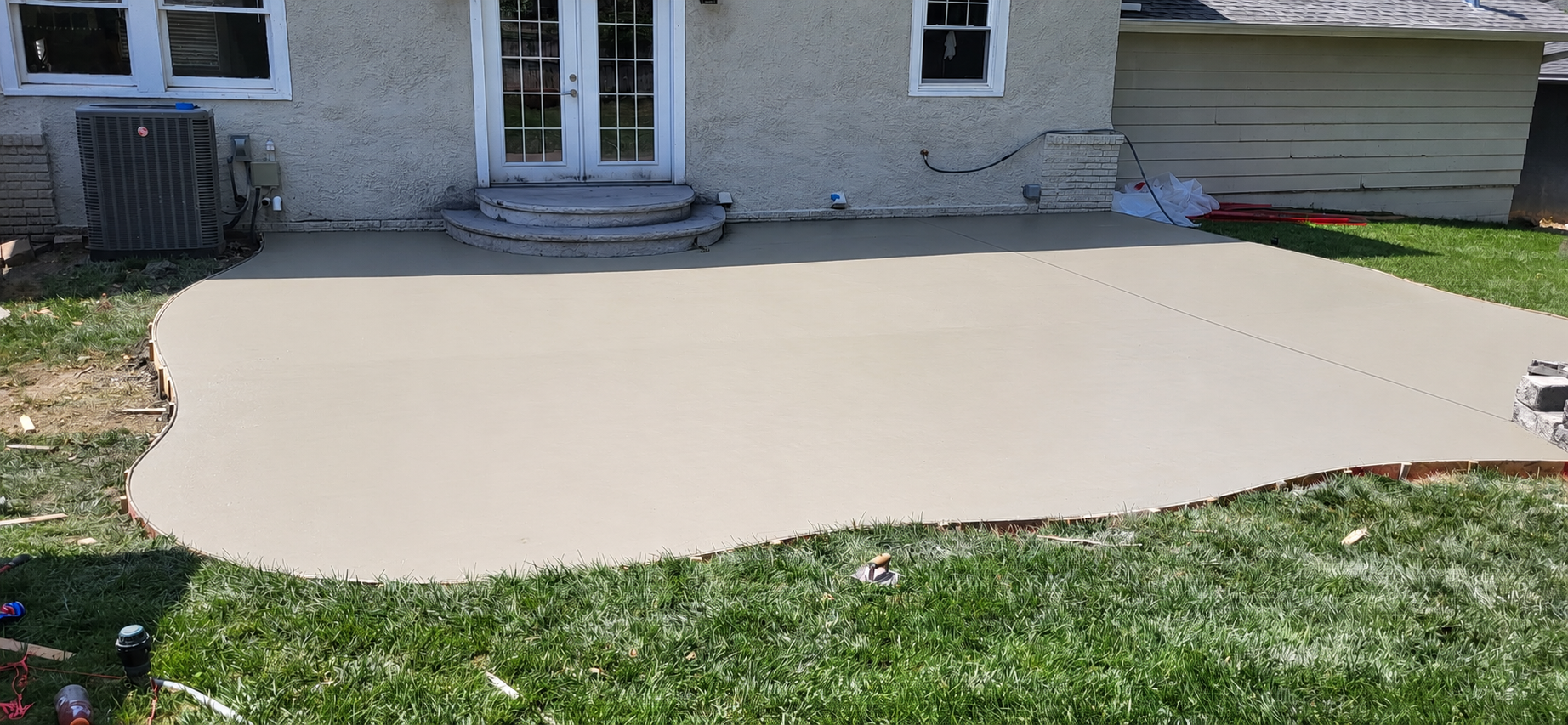 Freshly poured concrete patio in front of a house with steps leading to a glass double door. There's construction debris and tools around, with grass in the foreground.