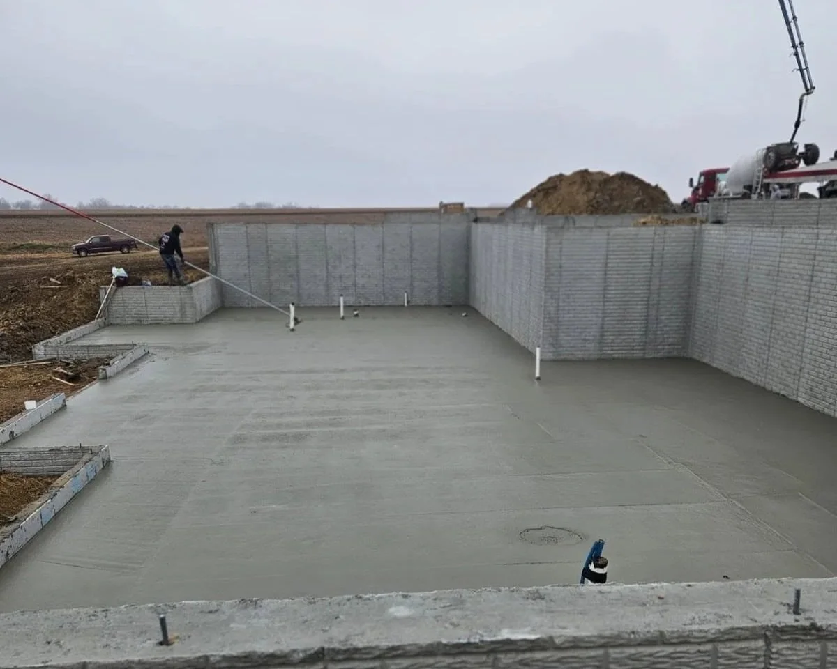 Construction site with a concrete foundation and perimeter walls, workers pouring concrete, and construction machinery visible on the site.