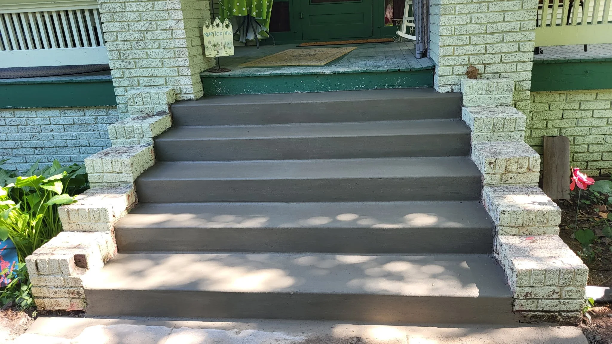 Concrete steps leading to a porch with a green door. The porch has a small welcome sign, a green curtain, and outdoor furniture. There is a flower on the right side of the stairs and greenery on the left.