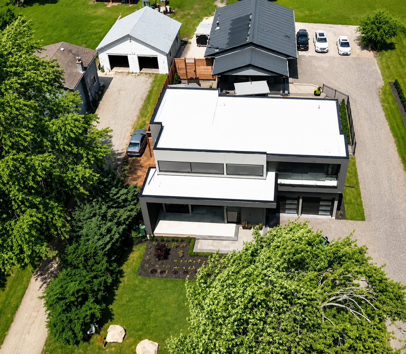 Aerial view of a modern house with a flat white roof, large windows, and a courtyard, surrounded by trees, neighboring houses, and a gravel driveway with parked cars.
