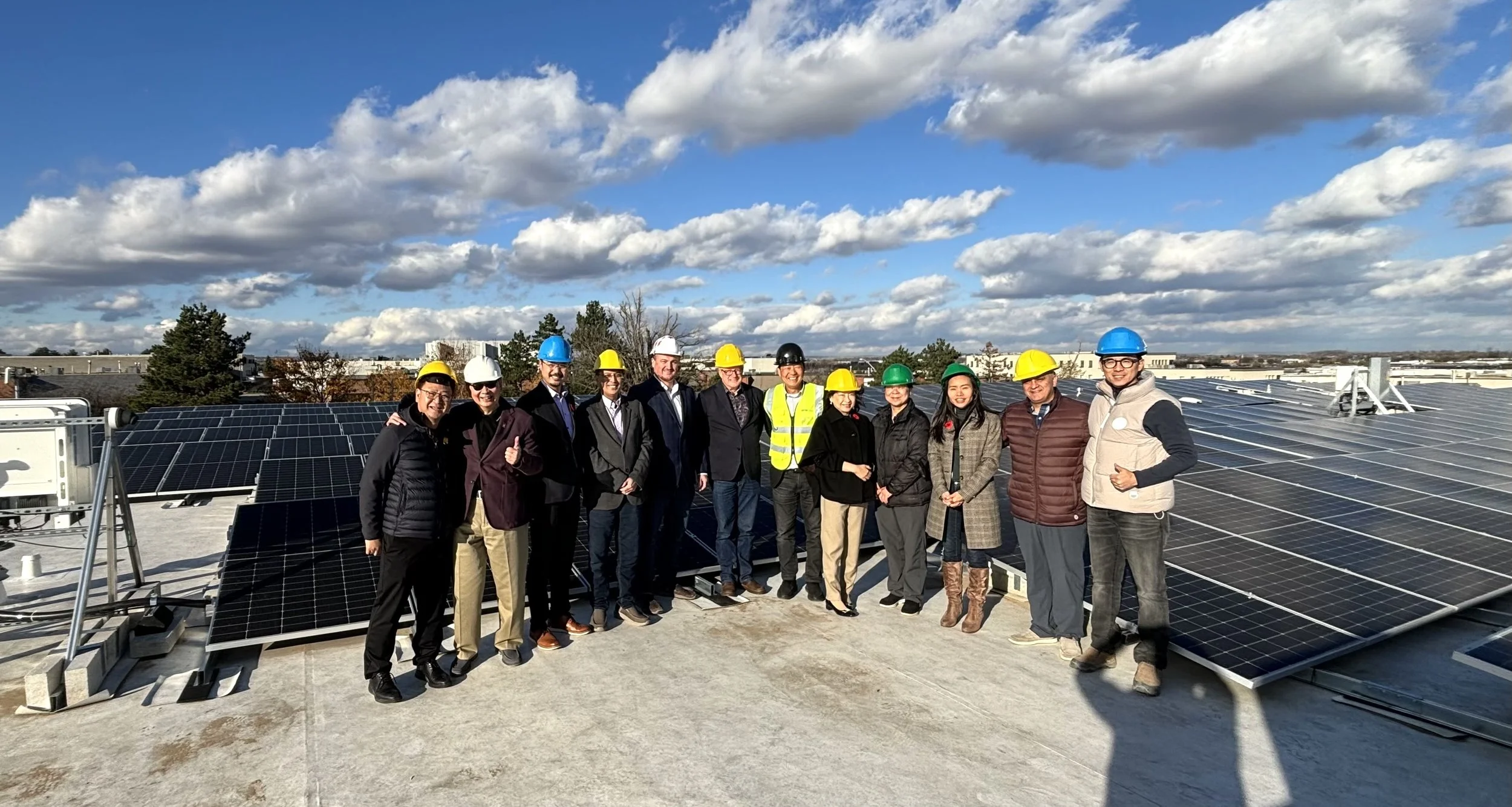 Group of people on a rooftop with solar panels, wearing safety helmets and jackets, under a partly cloudy sky.