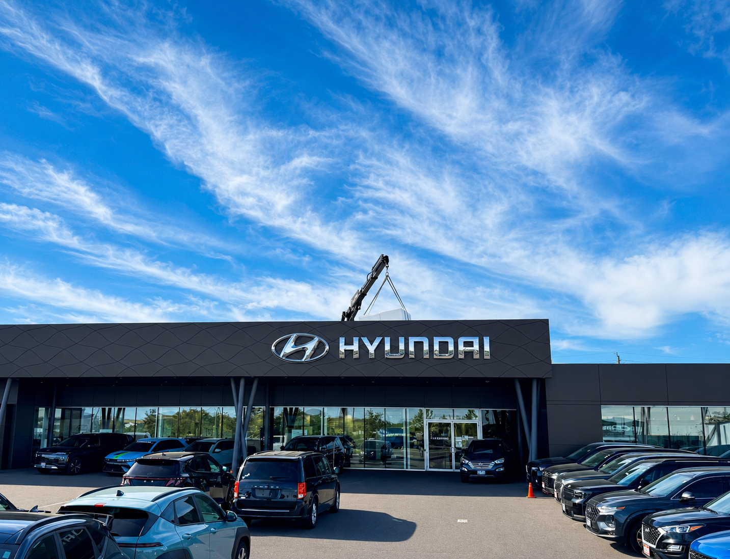 Hyundai dealership with a parking lot full of cars, a large Hyundai sign on the building, and a crane installing or repairing the sign, under a blue sky with scattered clouds.
