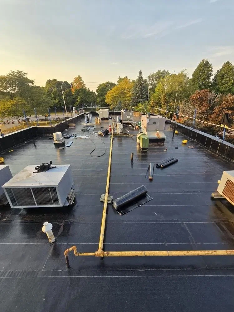 View of a building rooftop with HVAC units, pipes, and vents, surrounded by trees and a clear sky.