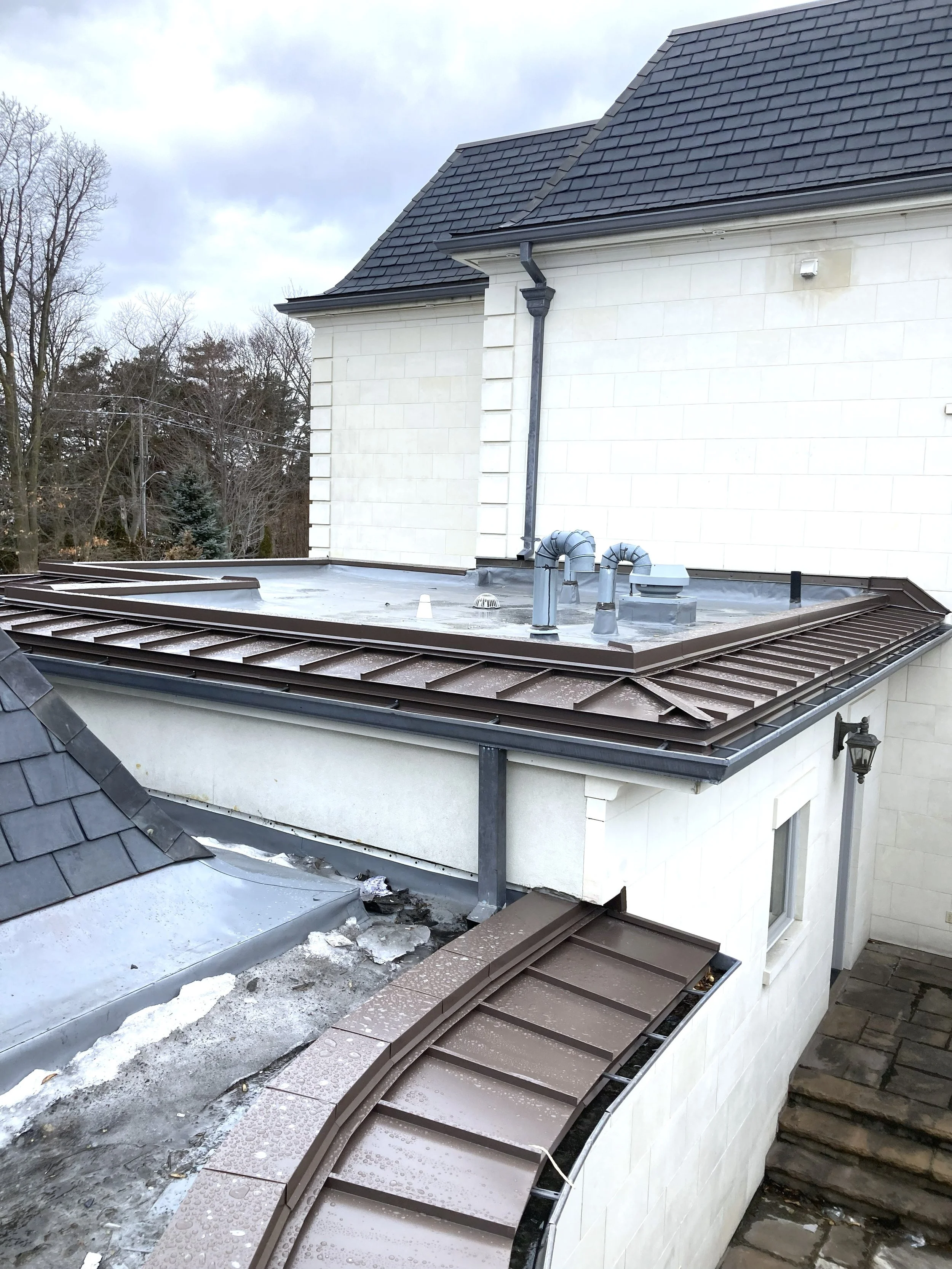 The image shows the roof of a house under construction or repair, with a flat, metal roofing surface and some tools and materials scattered around. The house has a sloped, shingled roof on the upper part of the image and a white exterior wall with a window and a wall-mounted lamp. There are trees in the background and the sky is partly cloudy.
