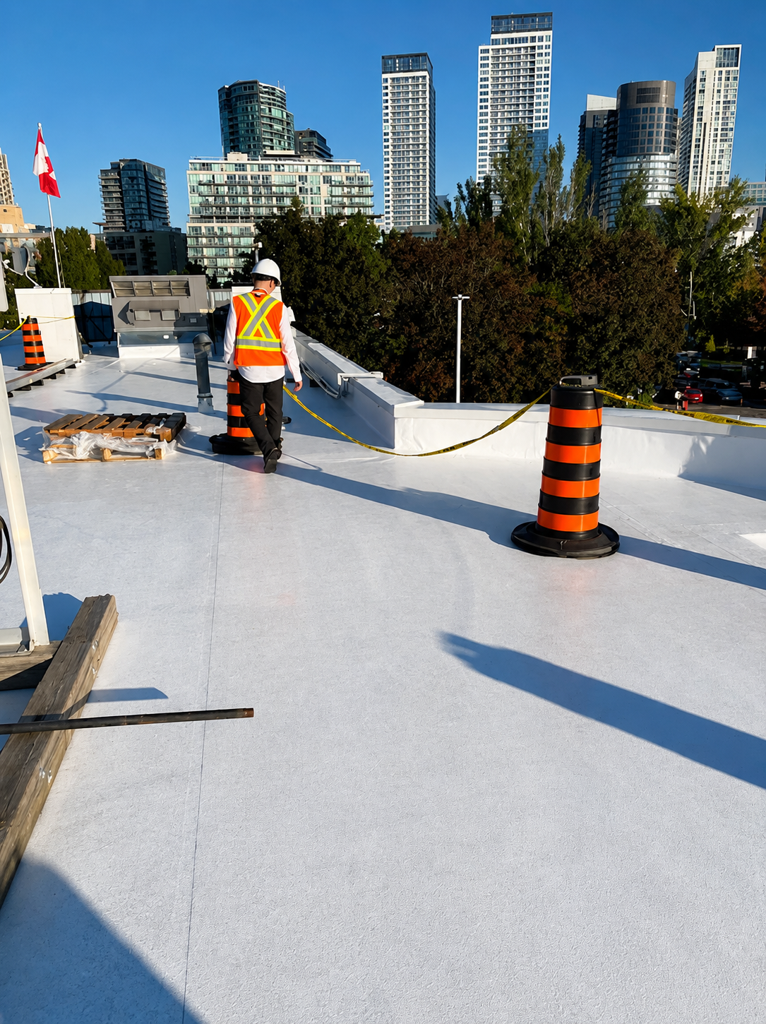 Construction worker on a rooftop with orange and black safety cones, trees, and city skyline in the background.