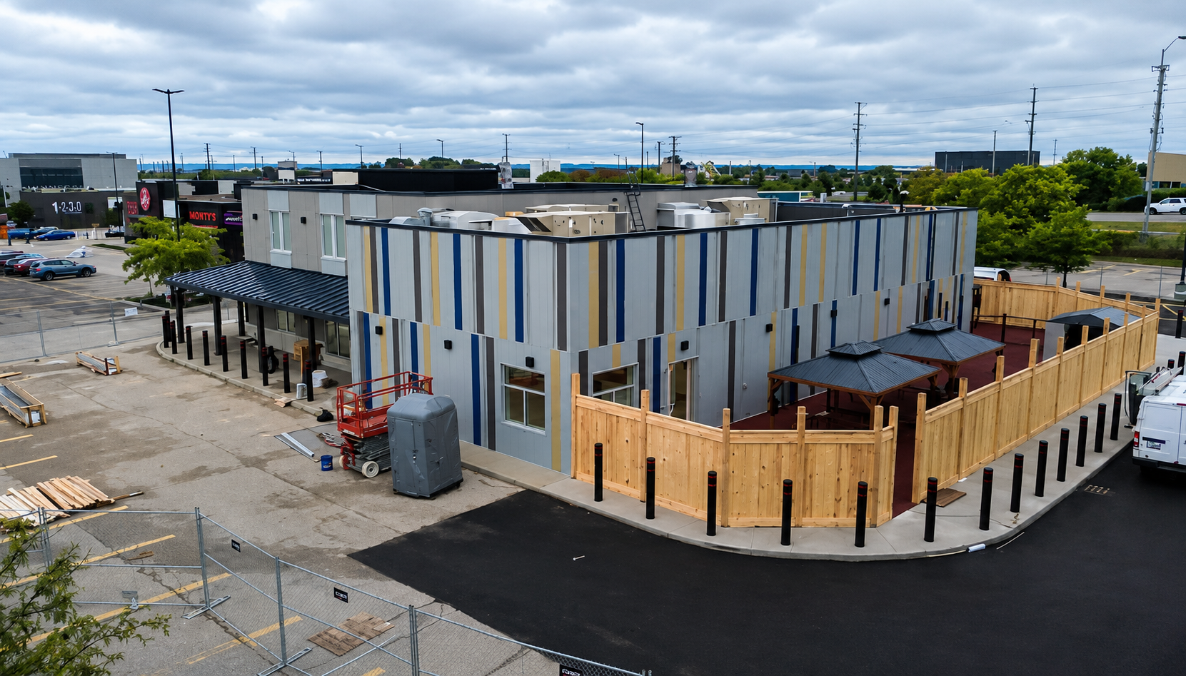 A newly constructed building with a colorful striped exterior, a fenced outdoor seating area with gazebos, and a parking lot with some construction materials nearby.