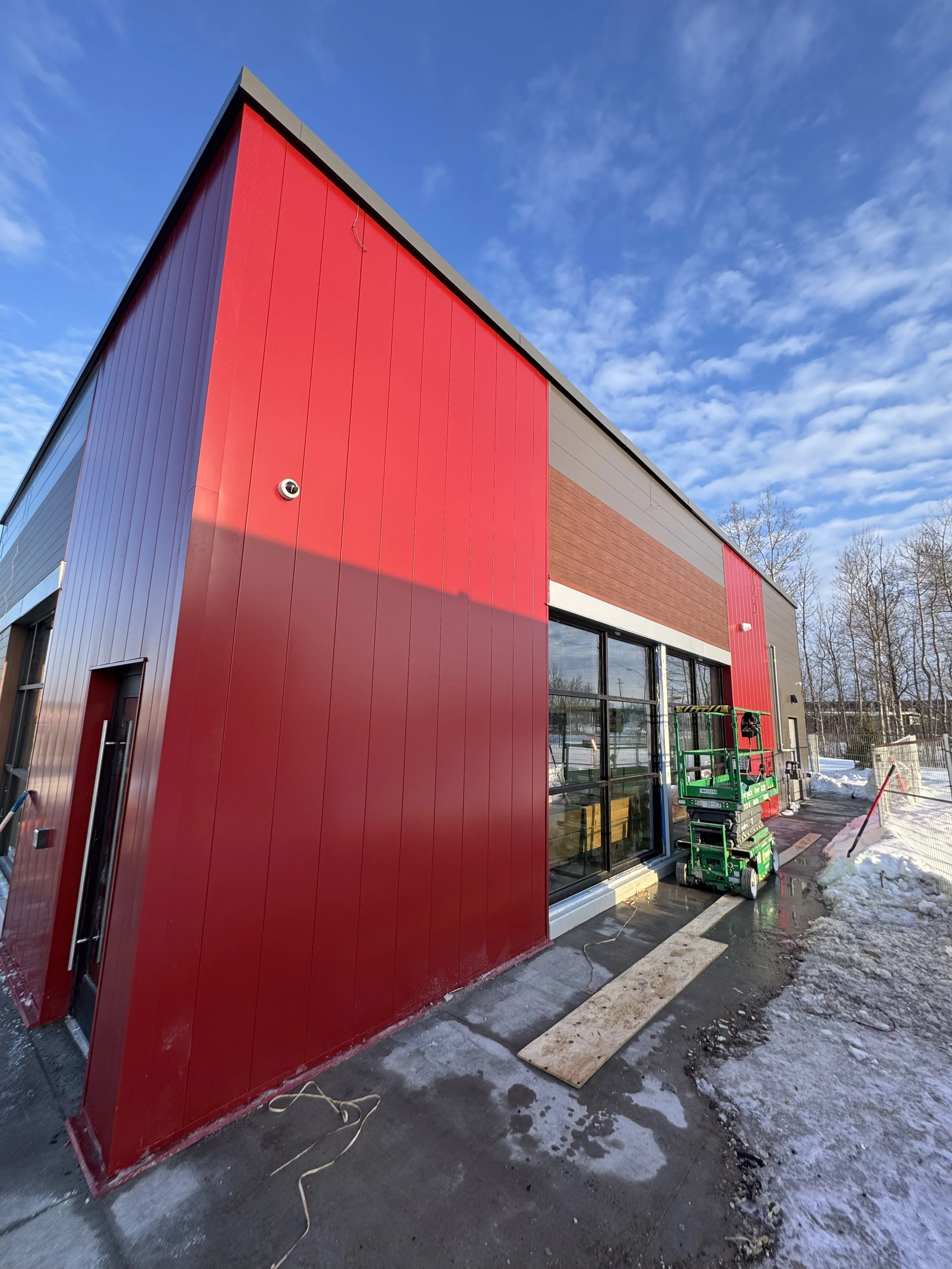 View of a modern building under construction with red and gray exterior panels, large windows, and construction equipment outside on a winter day with snow on the ground and a partly cloudy sky.