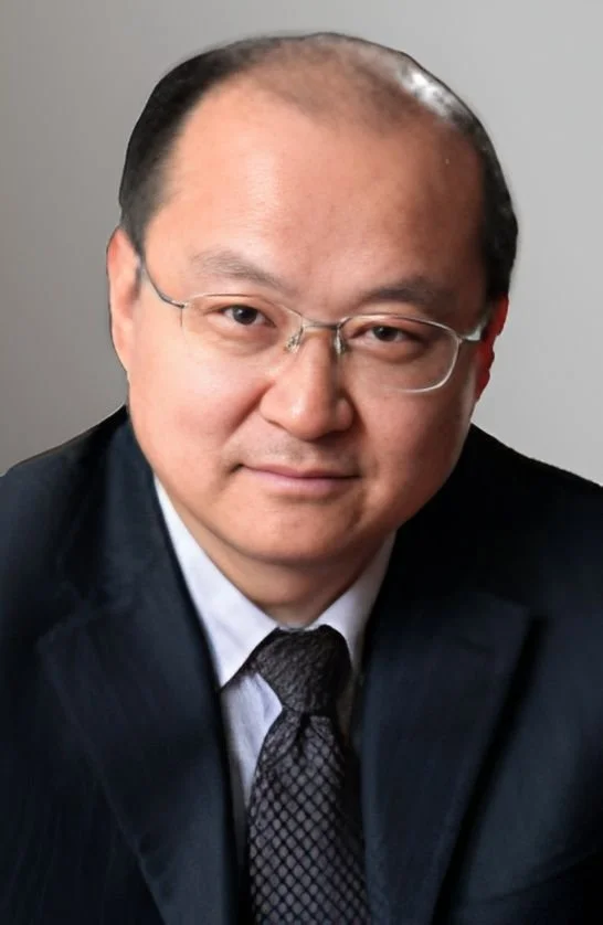 A man wearing glasses in a dark suit, white shirt, and patterned tie, facing the camera with a neutral expression, against a plain gray background.