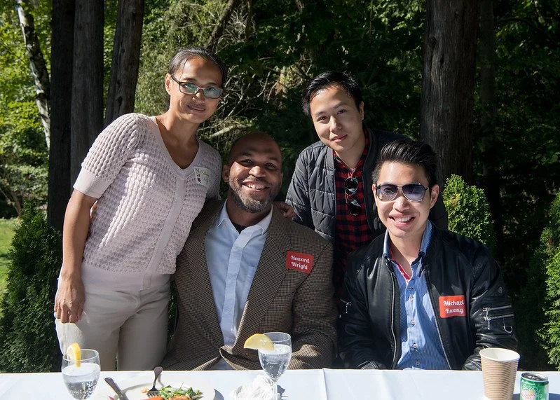 Four people smiling at a table outdoors, with three standing and one sitting, surrounded by trees and greenery.