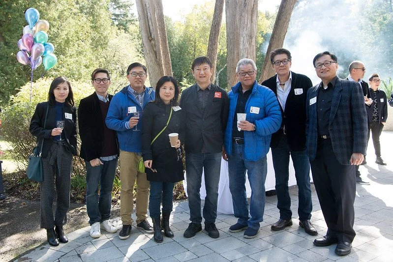 Group of eight people standing outdoors, posing for a photo during an event, some holding drinks, with trees and balloons in the background.