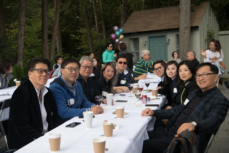 People sitting at a long outdoor table during a gathering or celebration, with a wooded background and some balloons.