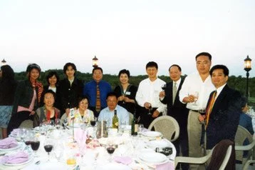 Group of people at a formal outdoor dinner, some holding glasses, with a white tablecloth, wine glasses, and dining ware in the foreground.