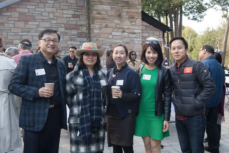 Five people standing outdoors in a social gathering, holding coffee cups, smiling, with other attendees in the background near a brick building and trees.