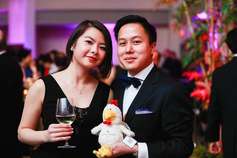 A young woman and a young man dressed in formal attire posing at a festive event. The woman holds a glass of white wine, and the man holds a plush toy of a duck wearing a chef's hat. The background features colorful decorations and other guests.