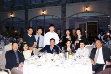 Group of people gathered around a table at a formal dinner event outside a building with large windows.